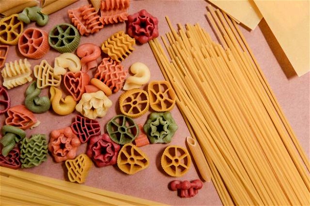 colorful pasta in different shapes on a pink surface, framed by piles of spaghetti
