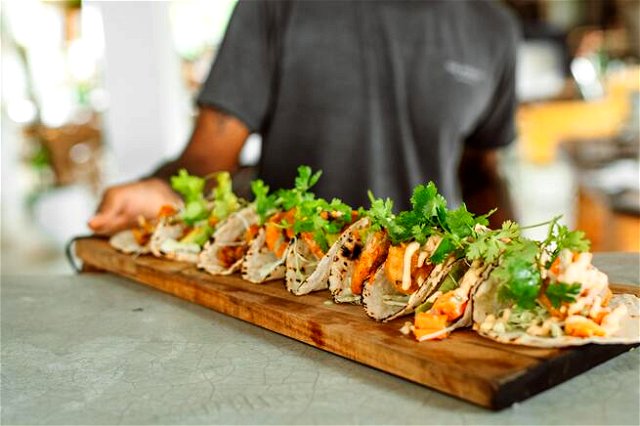 A person holding a wooden board with 9 tacos on it, colorful and covered in fresh herbs