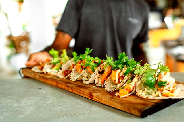 A person holding a wooden board with 9 tacos on it, colorful and covered in fresh herbs