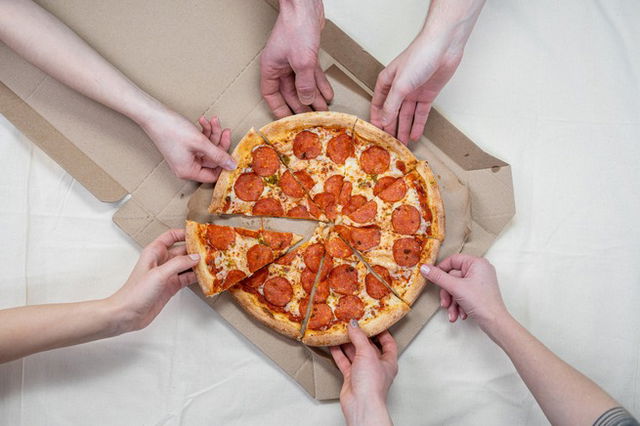 Pizza resting in a takeaway box, six arms each reaching for a slice from different directions.