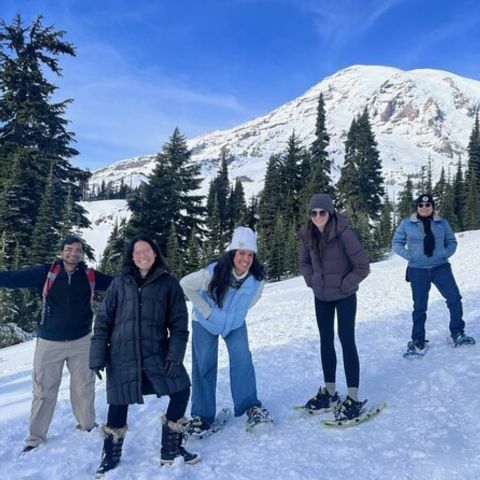 A group of people standing and snowshoeing on a snowy trail, smiling and posing in winter clothing, with tall evergreen trees and a large snow-covered mountain rising behind them under a clear blue sky.