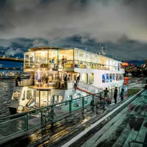 A brightly lit, multi-deck river cruise boat docked at night, with warm interior lights glowing through large windows, reflections shimmering on the water, and a wet wooden pier in the foreground under a cloudy sky.