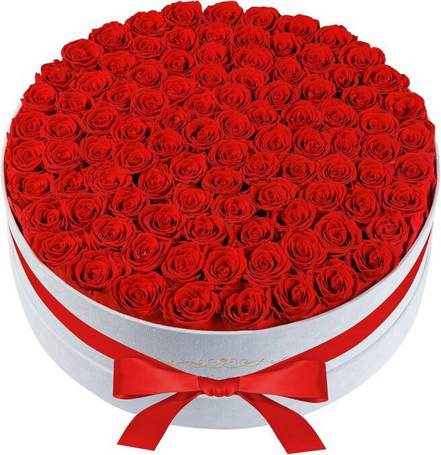 A round white gift box filled edge to edge with tightly arranged red roses, finished with a red satin ribbon tied in a bow around the base.