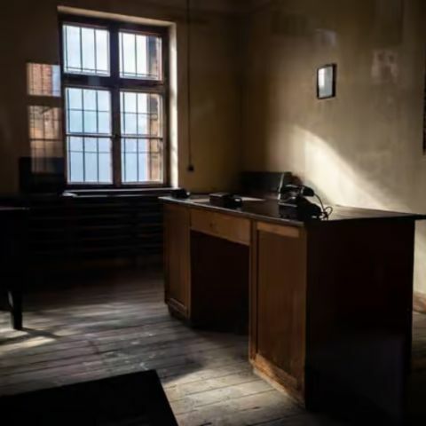 A dimly lit, sparsely furnished room with a wooden desk, chairs, and a barred window, sunlight casting long shadows across the worn floor, suggesting a historic interior space.