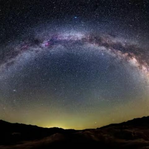 Wide view of the Milky Way arching across a dark night sky above a desert landscape with silhouetted hills.