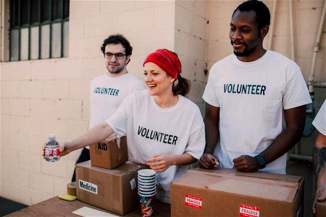 three people wearing teeshirts that say volunteers standing behind boxes handing out bottles, a female volunteer in a bandana is smiling.