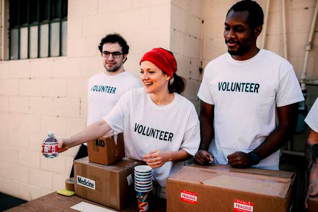 three people wearing teeshirts that say volunteers standing behind boxes handing out bottles, a female volunteer in a bandana is smiling.