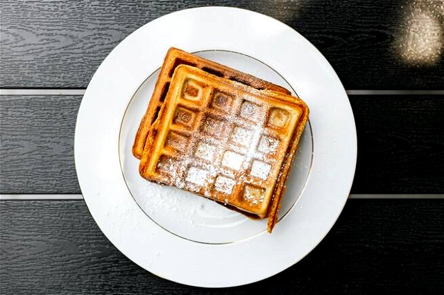 A waffle on a plate with a sprinkling of icing sugar. Served on a gold rimmed white plate on a black wooden boarded table.