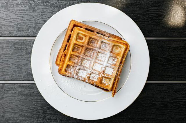 A waffle on a plate with a sprinkling of icing sugar. Served on a gold rimmed white plate on a black wooden boarded table.