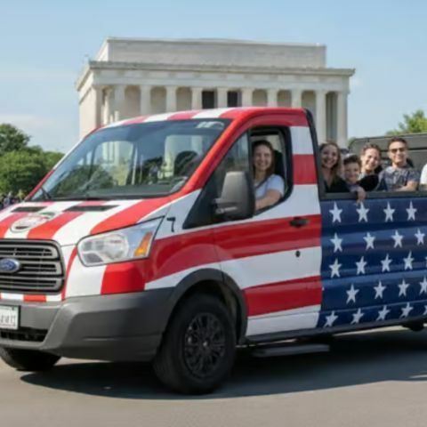 A group of smiling people riding in an open-sided tour van painted like the American flag, driving along a city street with a large white monument visible in the background on a sunny day.